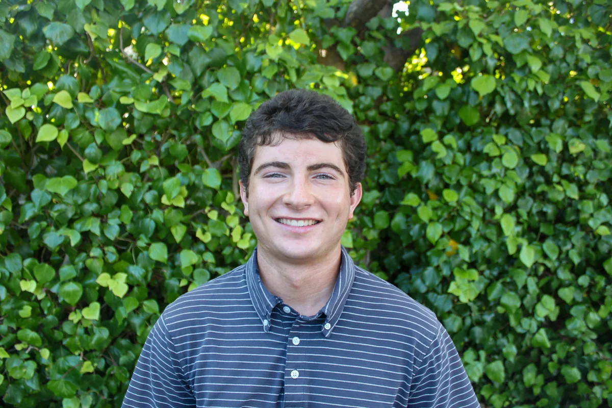 Portrait of Gus, smiling, in a striped shirt in front of an ivy-covered wall.
