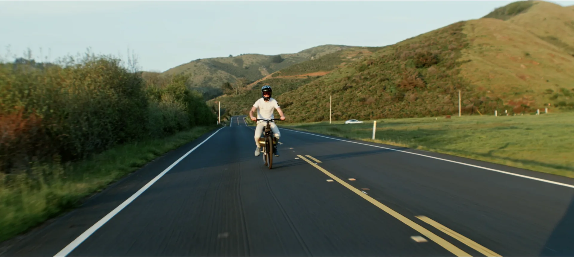 A rider on The Nacho from behind, heading up an empty road through green coastal hills.
